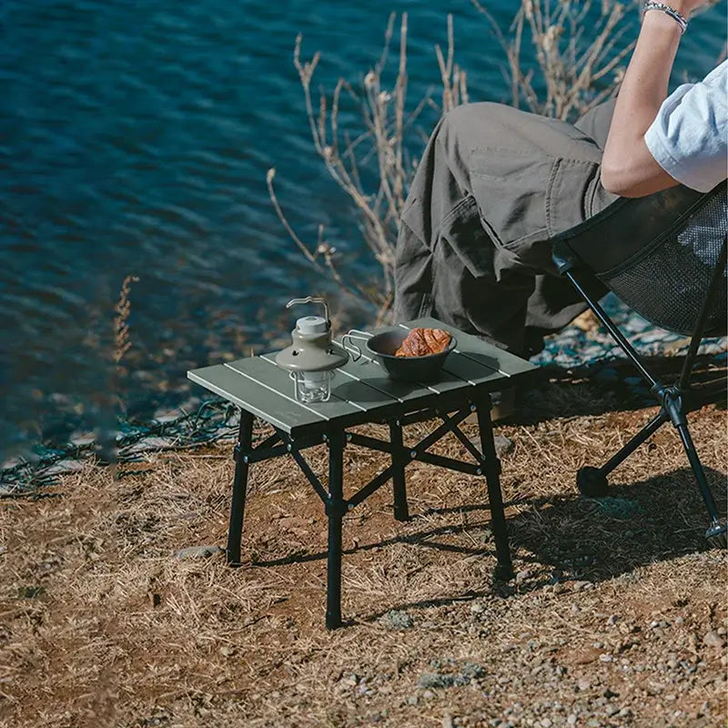 Portable aluminum alloy folding table set up for outdoor camping by the water, featuring a drink and snack.