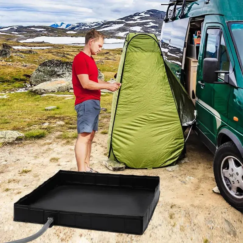 Man using a portable camping shower tent with a black floor base next to a green van in a scenic landscape.